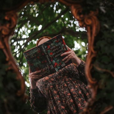 A girl with red hair and in a green dress with red flowers on it holds a green and red book titled Immortal Longings by Chloe Gong. The person is reflected in a bronze ornate mirror with leaves surrounding the mirror.